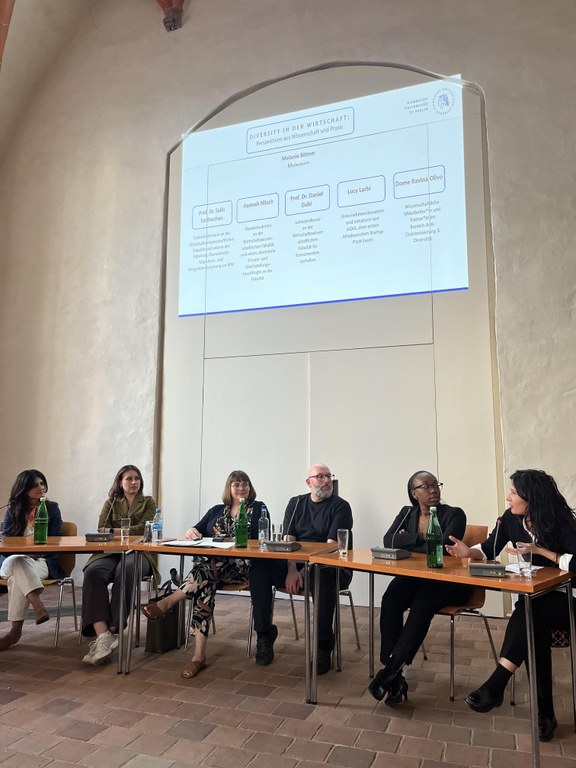 The photo shows the panellists of the panel discussion "Diversity in Business: Perspectives from Science and Practice" on July 5th, 2023. From left to right: Prof. Dr. Sulin Sardoschau, Hannah Nitsch, Melanie Bittner, Prof. Dr. Daniel Guhl, Lucy Larbi and Dome Ravina Olivo. A powerpoint slide is projected on the wall above the panelists with the title of the discussion and the names (with their areas of expertise) in the seating order.